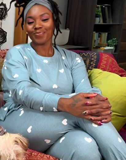 Woman wearing a teal blue pajama set with heart patterns, standing in a bedroom with tufted headboard.