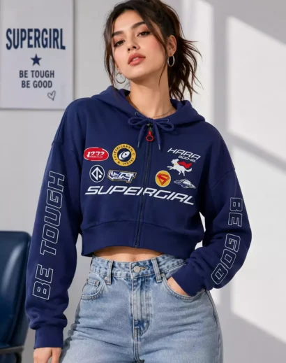 Women's Supergirl Blue Cropped Jacket front view with graphic patches and zip-up hoodie design paired with light blue denim jeans.
