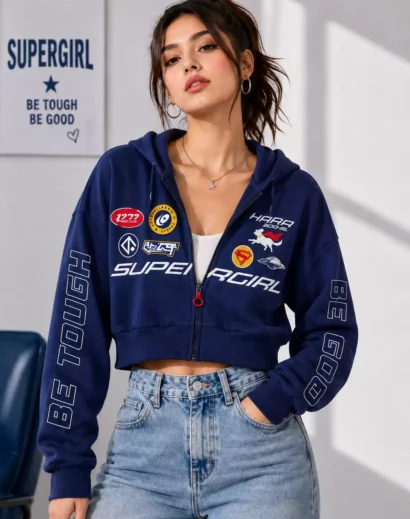 Women's Supergirl Blue Cropped Jacket with half zip open styling, graphic patches and cropped hoodie paired with denim jeans.